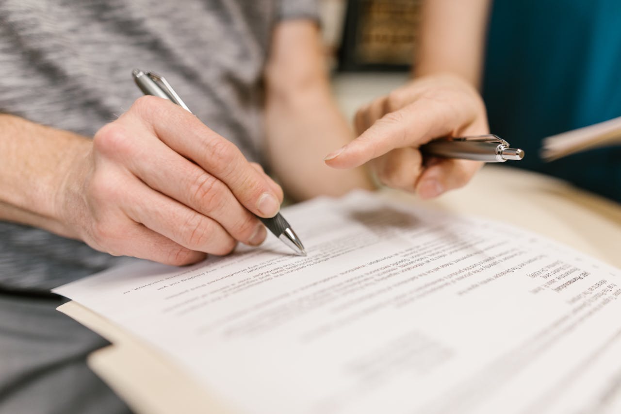 Close-up of two adults reviewing and signing a legal document with pens indoors.